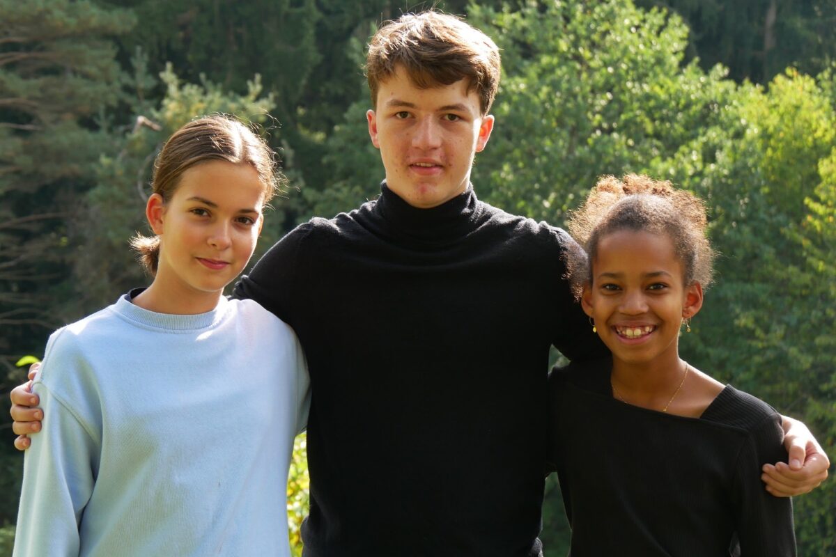 Drei Jugendliche stehen Arm in Arm lächelnd vor grünem Wald im Sonnenlicht