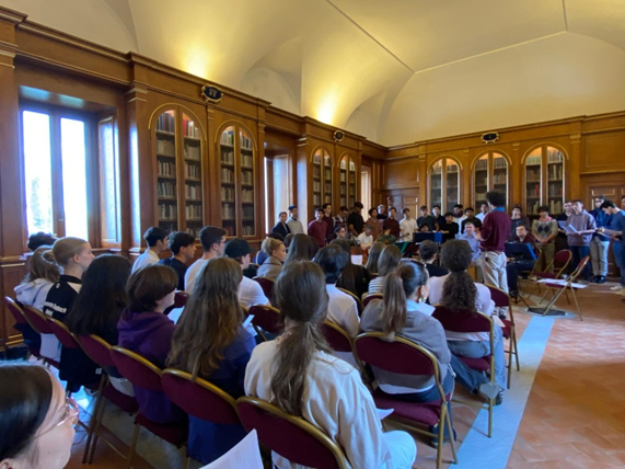 Viele junge Erwachsene sitzen aufmerksam in einem hellen Raum mit hohen Bücherregalen