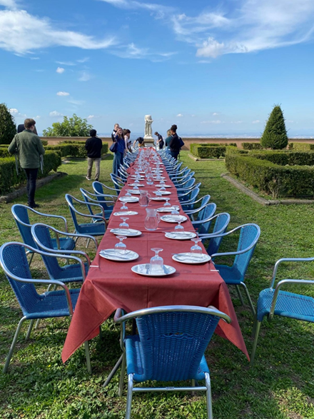Eine lange Tafel mit roten Tischtüchern im Garten, blaue Stühle, Menschen im Hintergrund, sonniges Wetter und eine Statue am Tischende