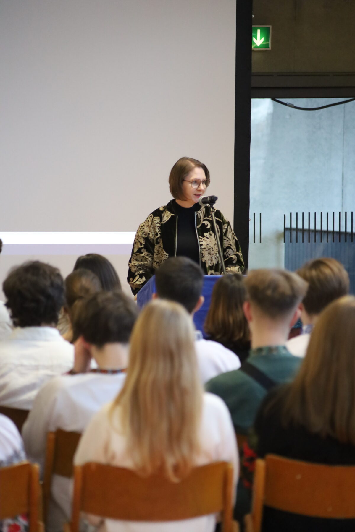 Eine Frau mit kurzem braunen Haar spricht lächelnd am Rednerpult vor Publikum