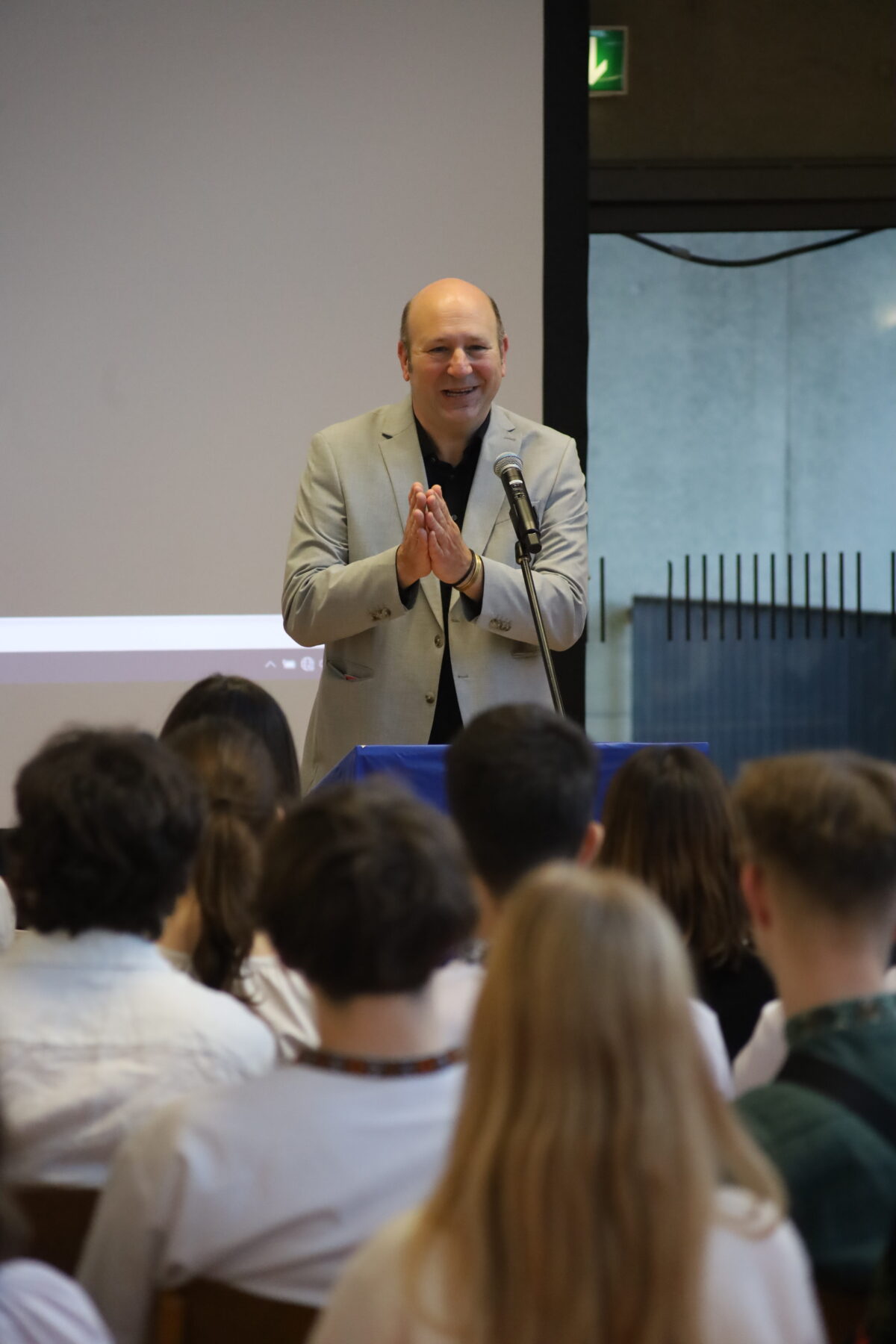 Ein glatzköpfiger Mann in hellem Sakko lächelt freundlich am Rednerpult vor Publikum