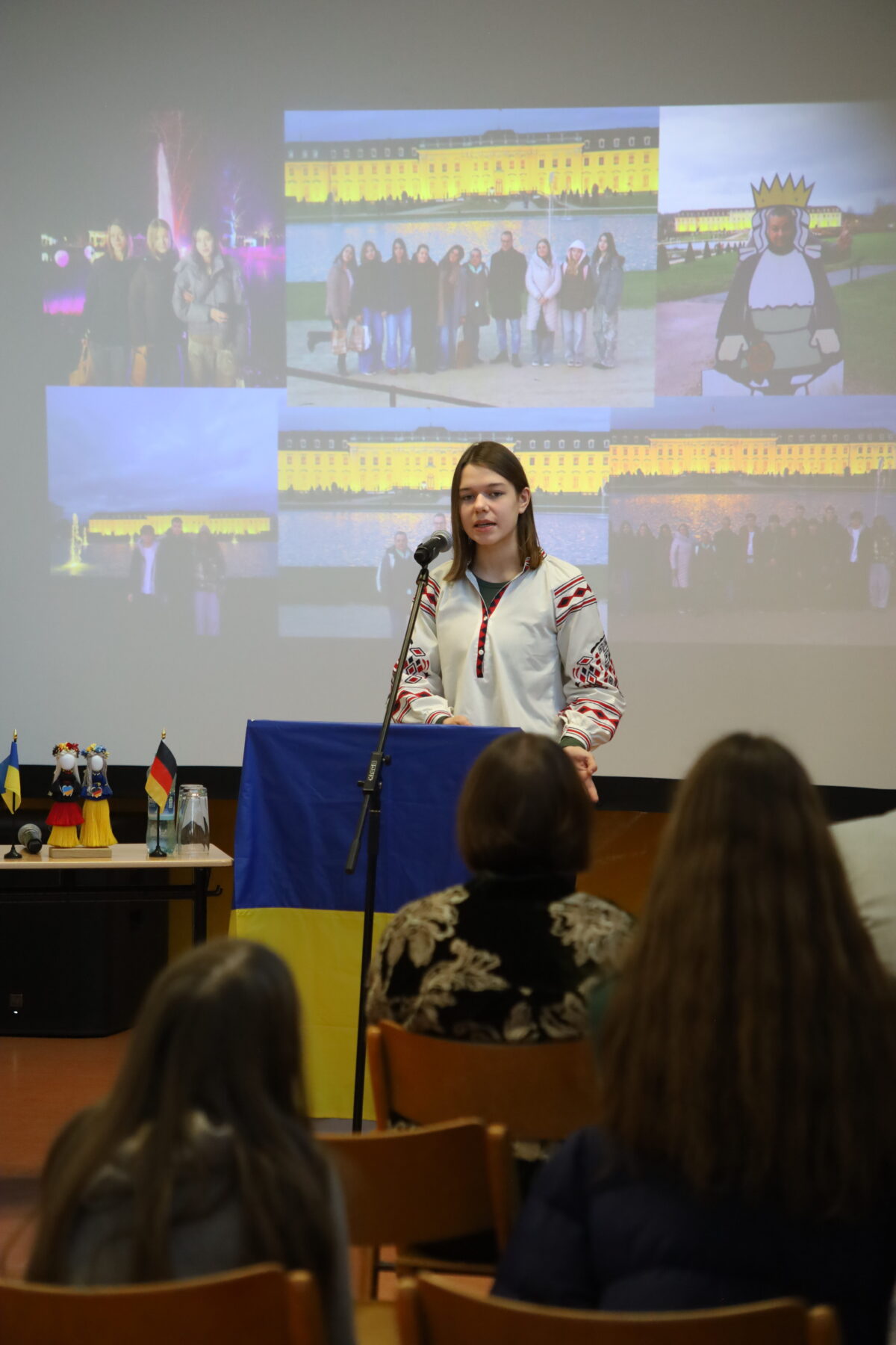 Junge Frau mit ernster Miene spricht am Rednerpult vor projizierten Gruppenfotos