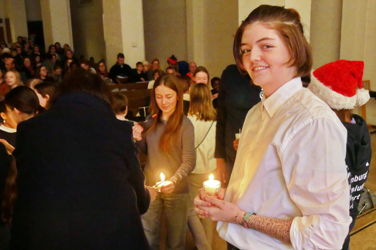 Eine junge Person mit hellem Hemd und kurzem Haar lächelt freundlich mit Kerze in der Hand