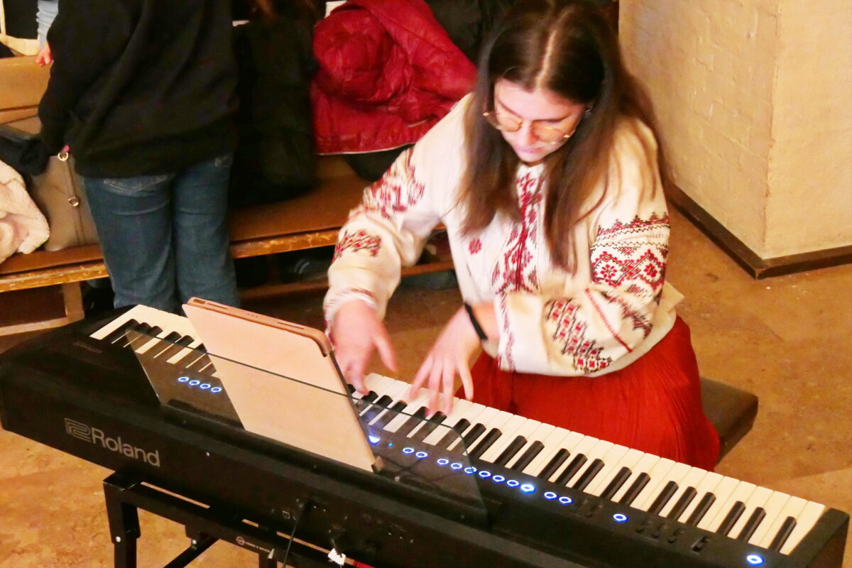 Eine Frau mit langen braunen Haaren spielt konzentriert Keyboard und trägt ein cremefarbenes Hemd mit rotem Muster