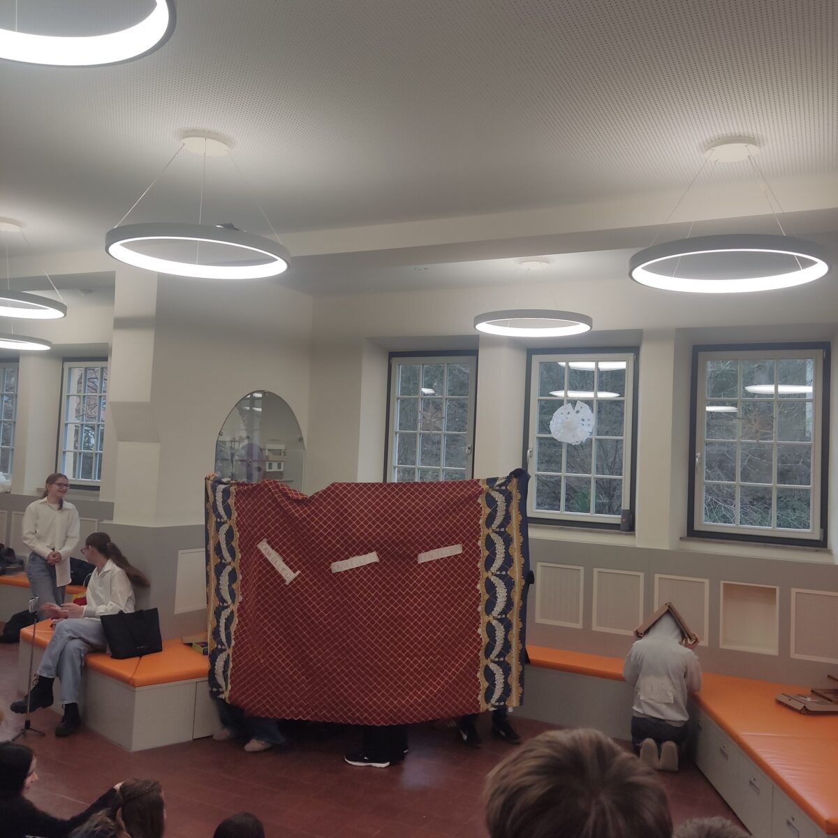 Mehrere Personen sitzen in einem hellen Raum, eine Gruppe hält einen gemusterten Teppich hoch