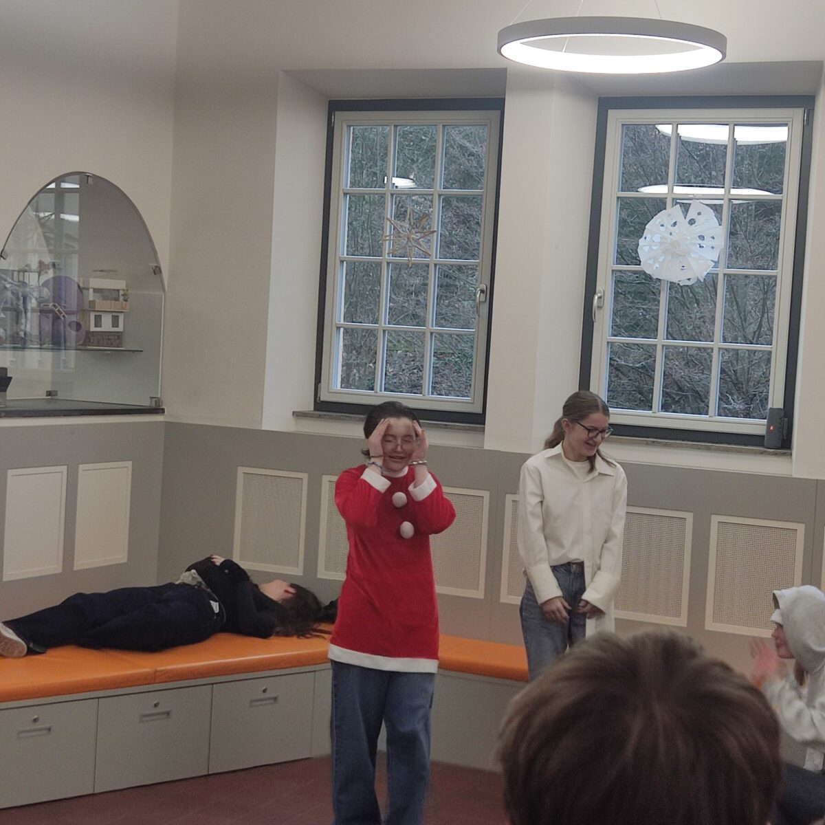 Eine lachende Person in rotem Kostüm steht vor Fenstern, während andere zuschauen