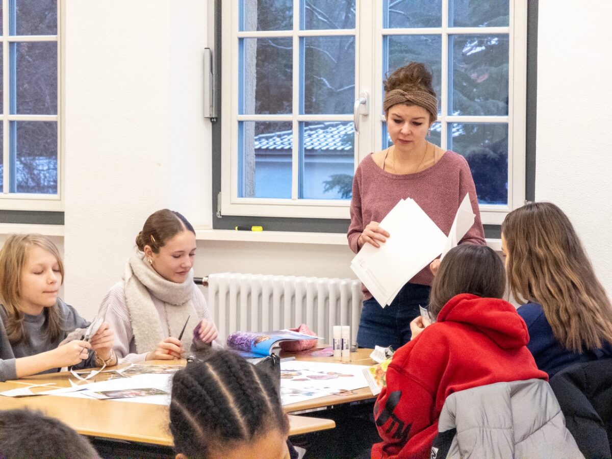 Eine Frau mit braunem Haar und Stirnband lächelt freundlich in einer Klasse