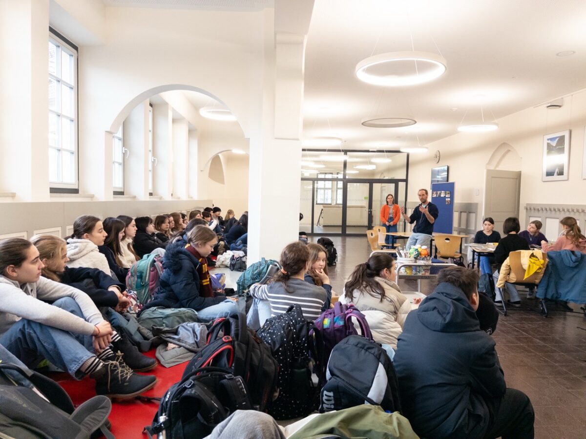 Gruppe junger Menschen sitzt aufmerksam in einem hellen Raum mit großen Fenstern