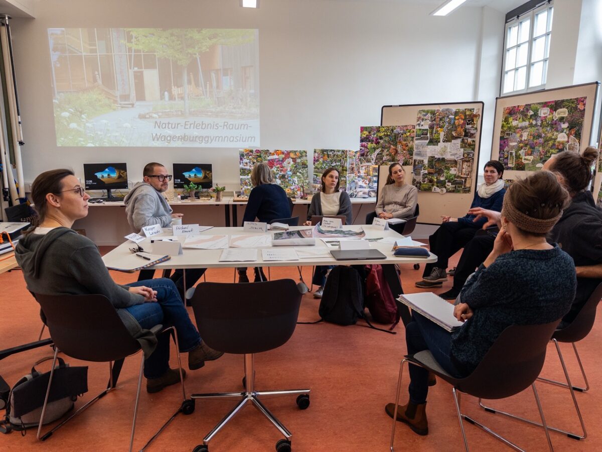 Mehrere Personen sitzen entspannt im Kreis in einem hellen Raum mit Projektor und bunten Plakaten