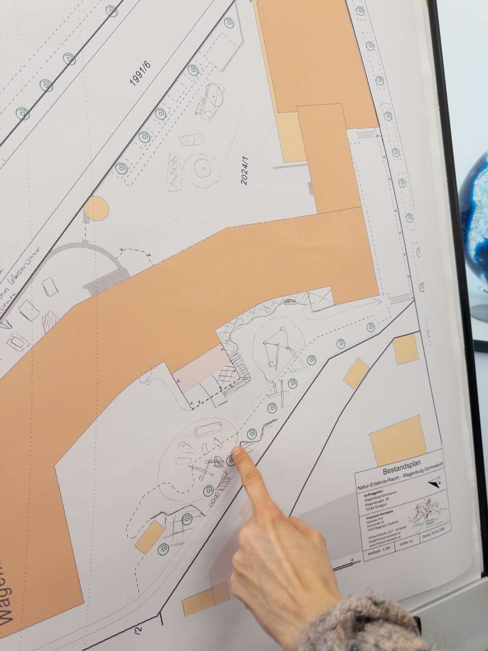 Eine Hand mit hellem Ärmel zeigt auf einen Punkt auf einem großen Architekturplan