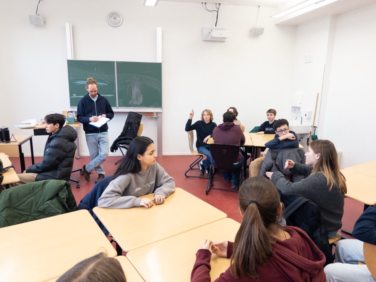 Ein Lehrer steht mit Papieren vor einer Tafel, Schüler diskutieren an Holztischen in einem hellen Klassenraum