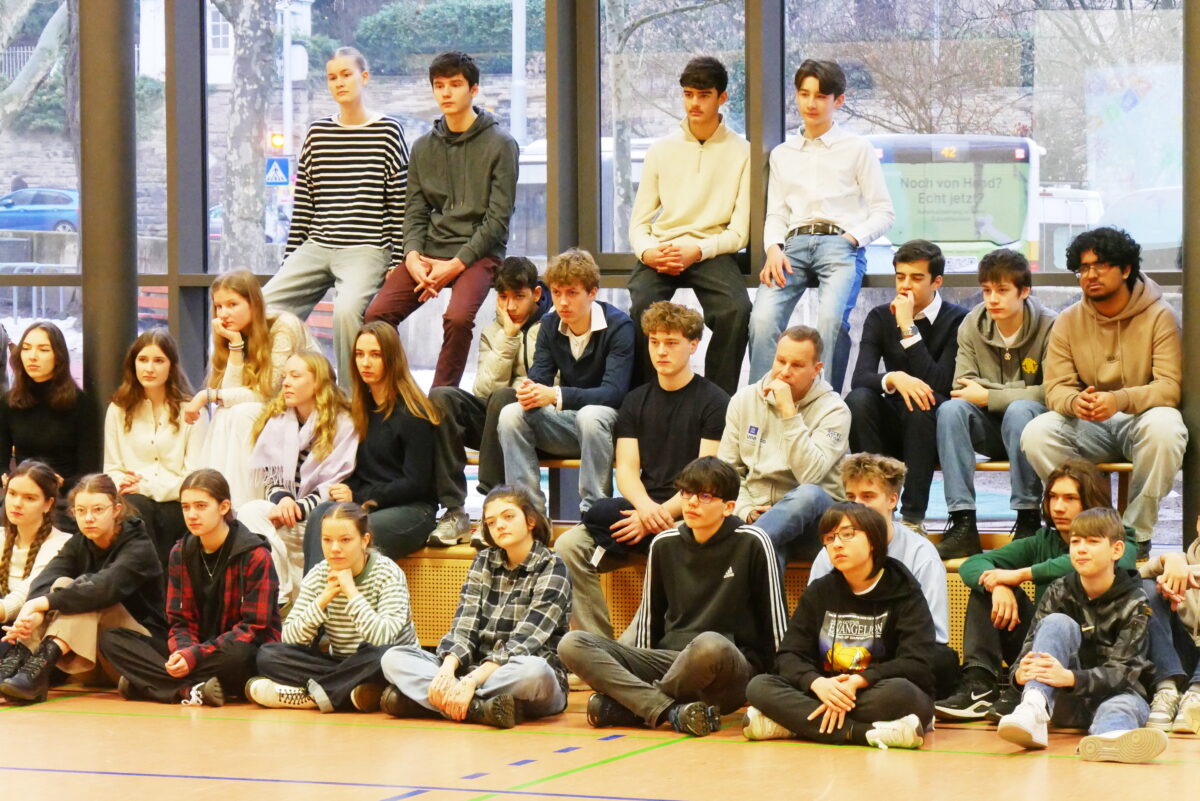 Eine Gruppe Jugendlicher sitzt entspannt in bunter Freizeitkleidung in einer Sporthalle