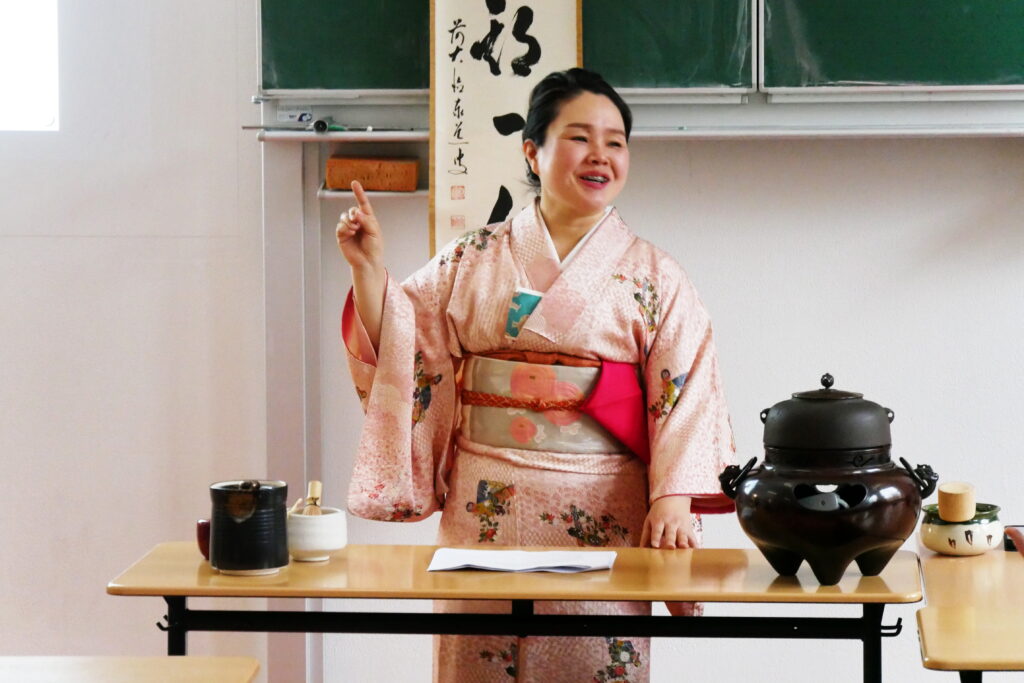 Eine asiatische Frau im rosa Kimono lächelt und hält einen Vortrag vor Teeservice