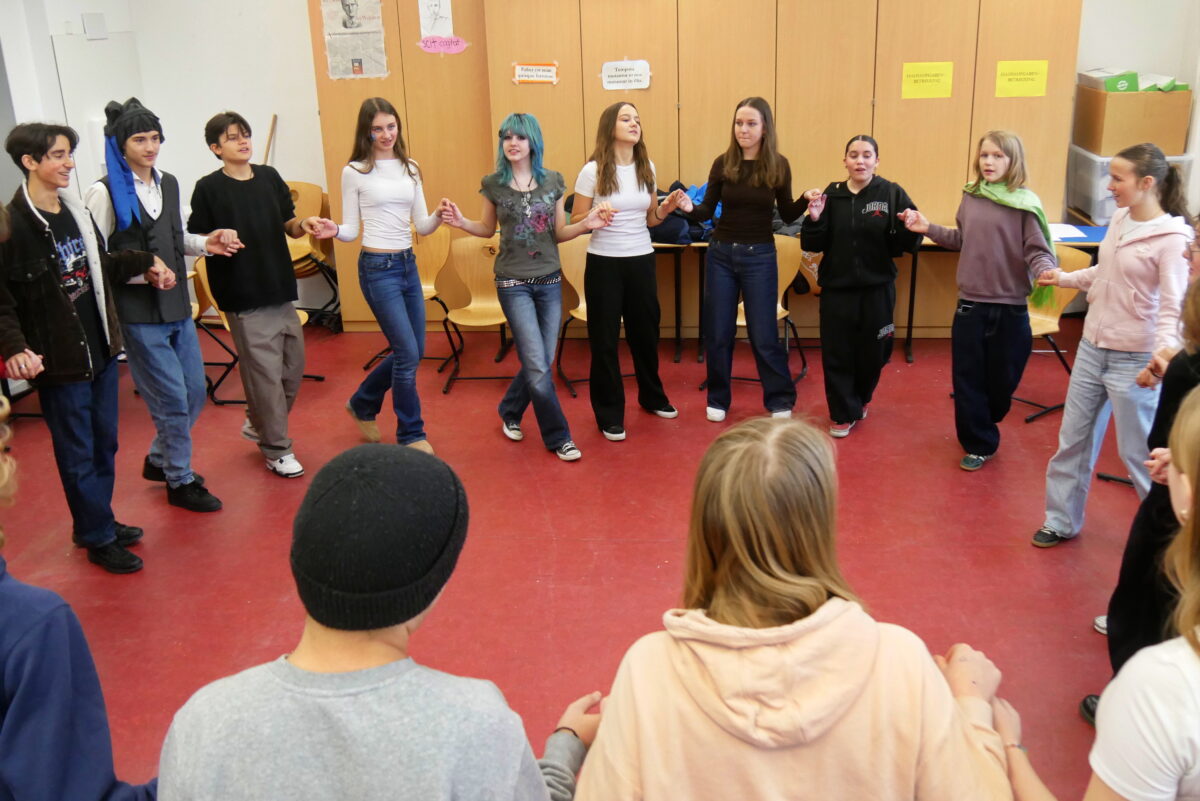 Eine Gruppe Jugendlicher tanzt lachend im Kreis auf einem rotbraunen Boden