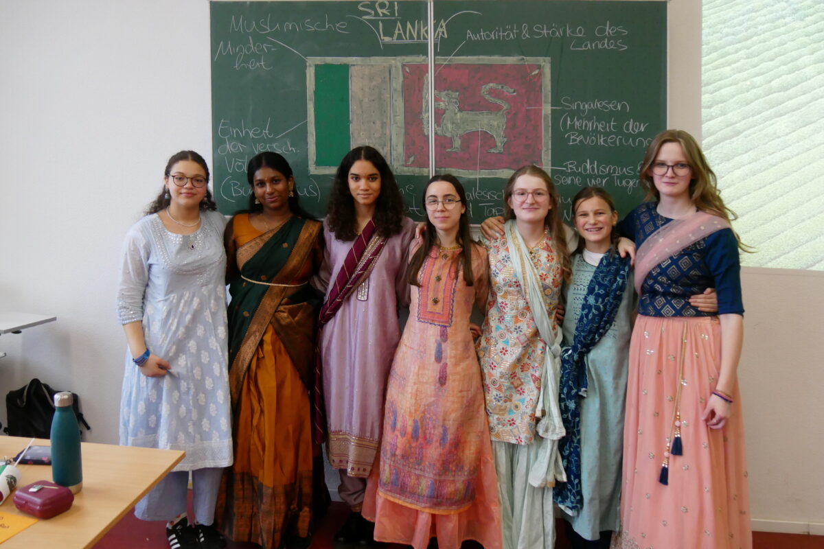 Sieben junge Frauen in bunter traditioneller Kleidung stehen lächelnd vor einer Tafel