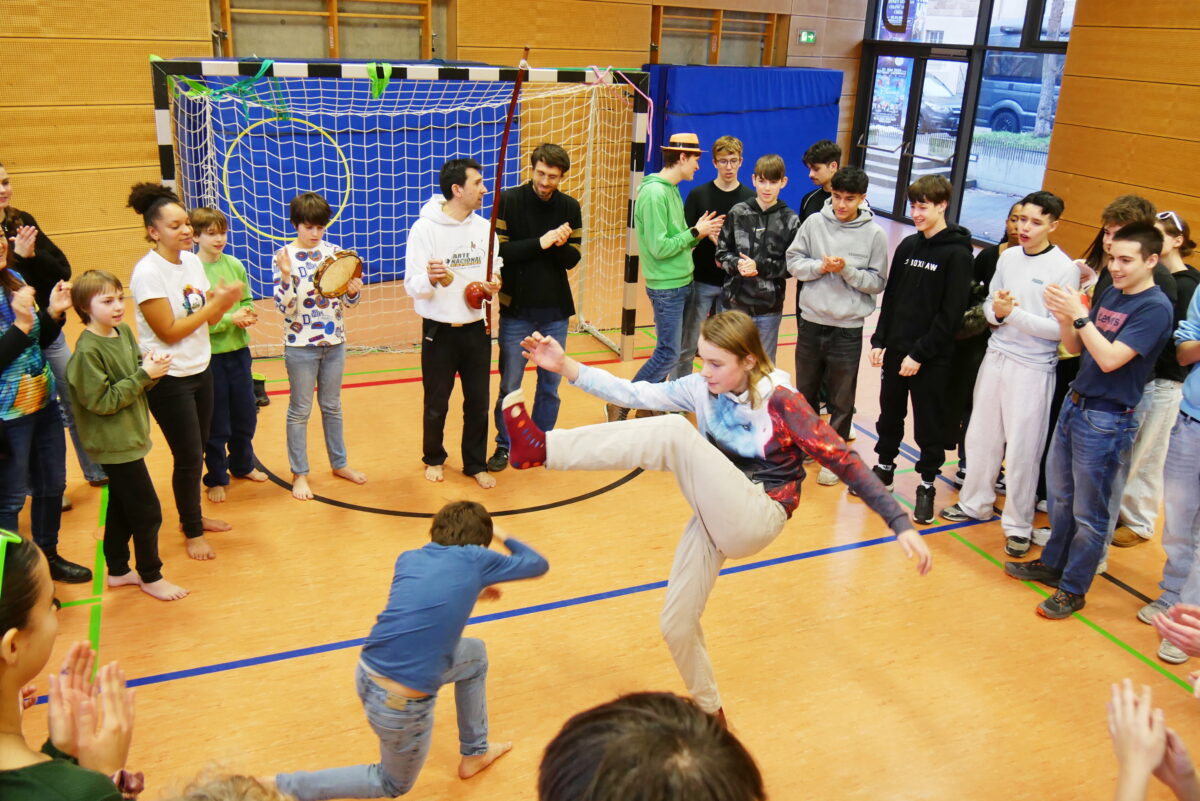 Eine Gruppe Jugendlicher steht lachend in einer Sporthalle, zwei davon tanzen Capoeira