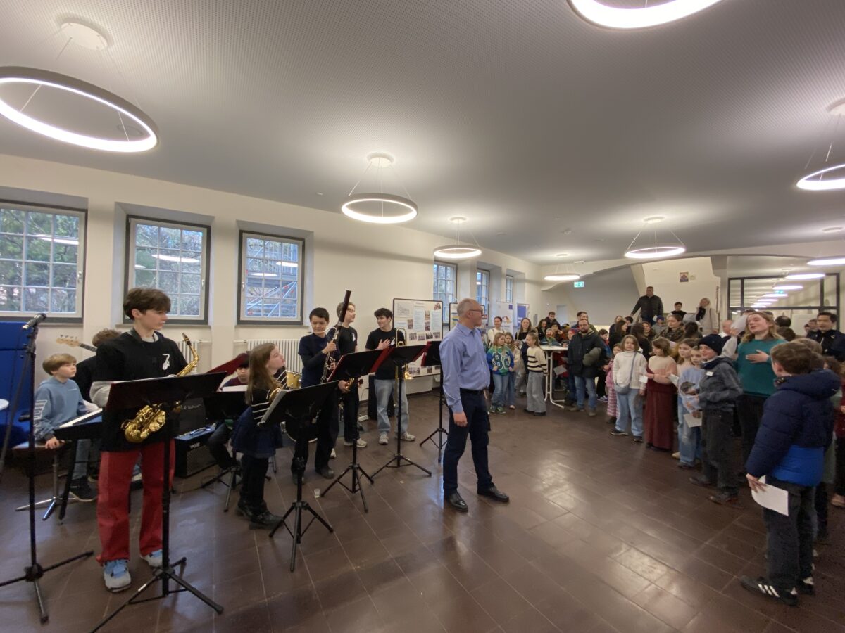 Eine Gruppe von Kindern spielt fröhlich Instrumente vor einem Publikum in heller Aula