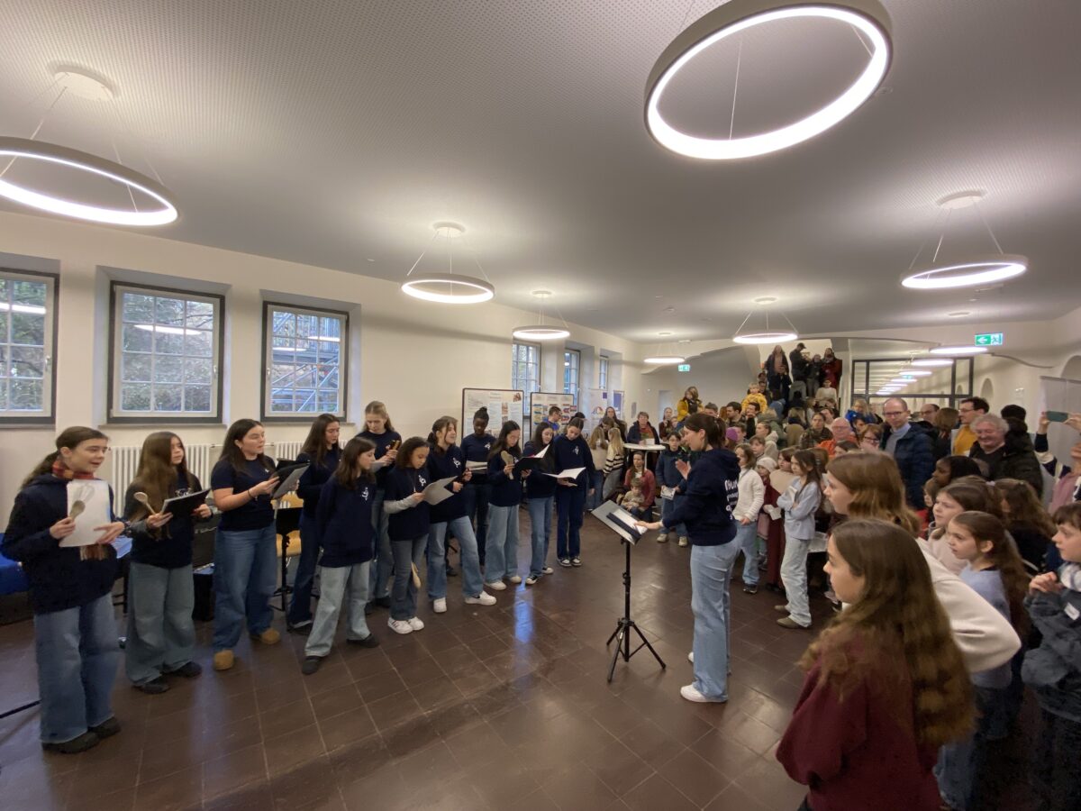 Eine Gruppe singender junger Frauen in blauen Pullovern steht fröhlich vor Publikum in einem hellen Raum mit runden Deckenleuchten