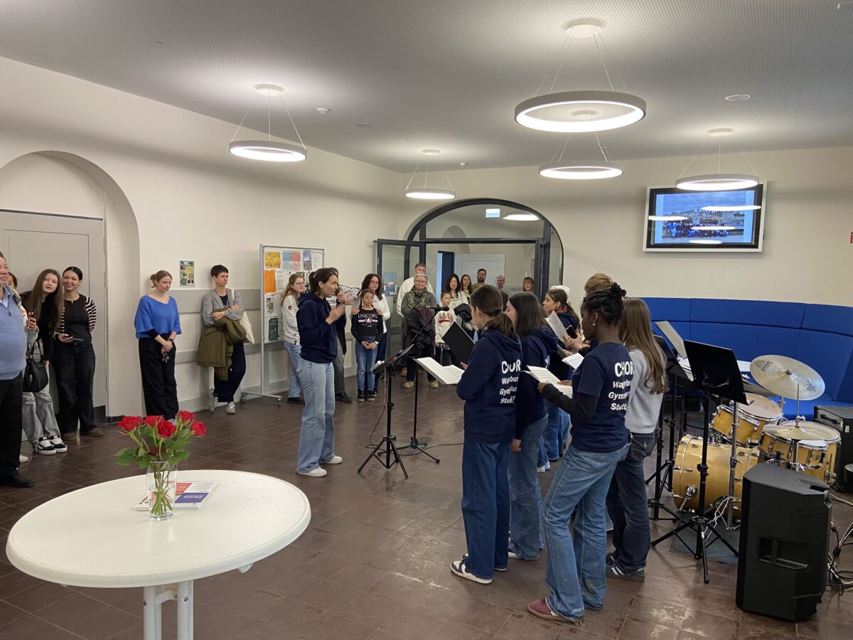 Eine Gruppe junger Menschen singt aufgeregt in einem hellen Raum vor interessiertem Publikum