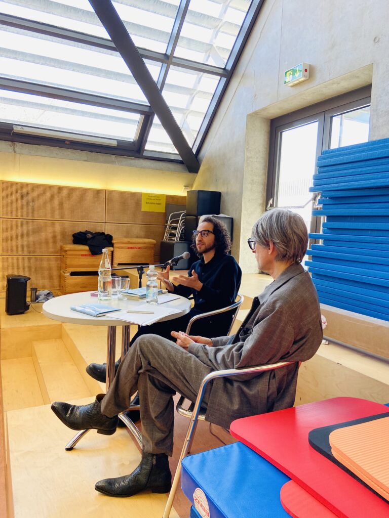 Zwei Personen sitzen an einem Tisch, einer spricht energiegeladen, beide in legerer Kleidung