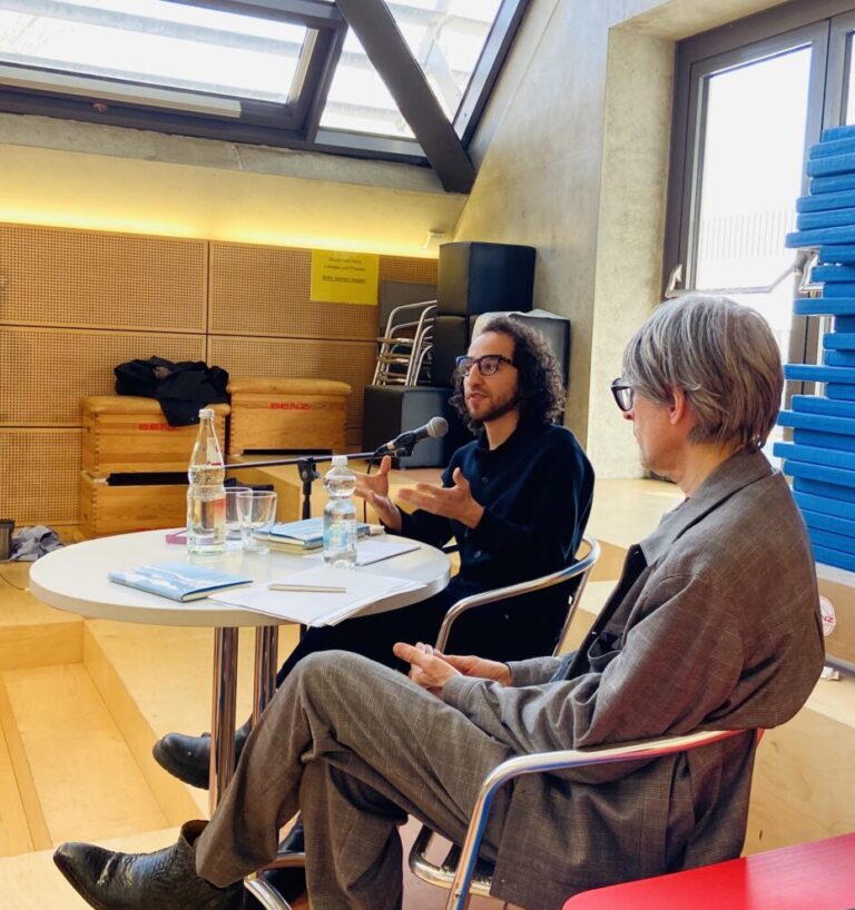 Zwei Personen sitzen an einem Tisch, einer spricht energiegeladen, beide in legerer Kleidung