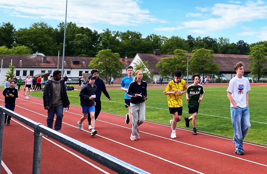 Gruppe joggt konzentriert auf roter Laufbahn neben gruenem Feld, bunte Kleidung
