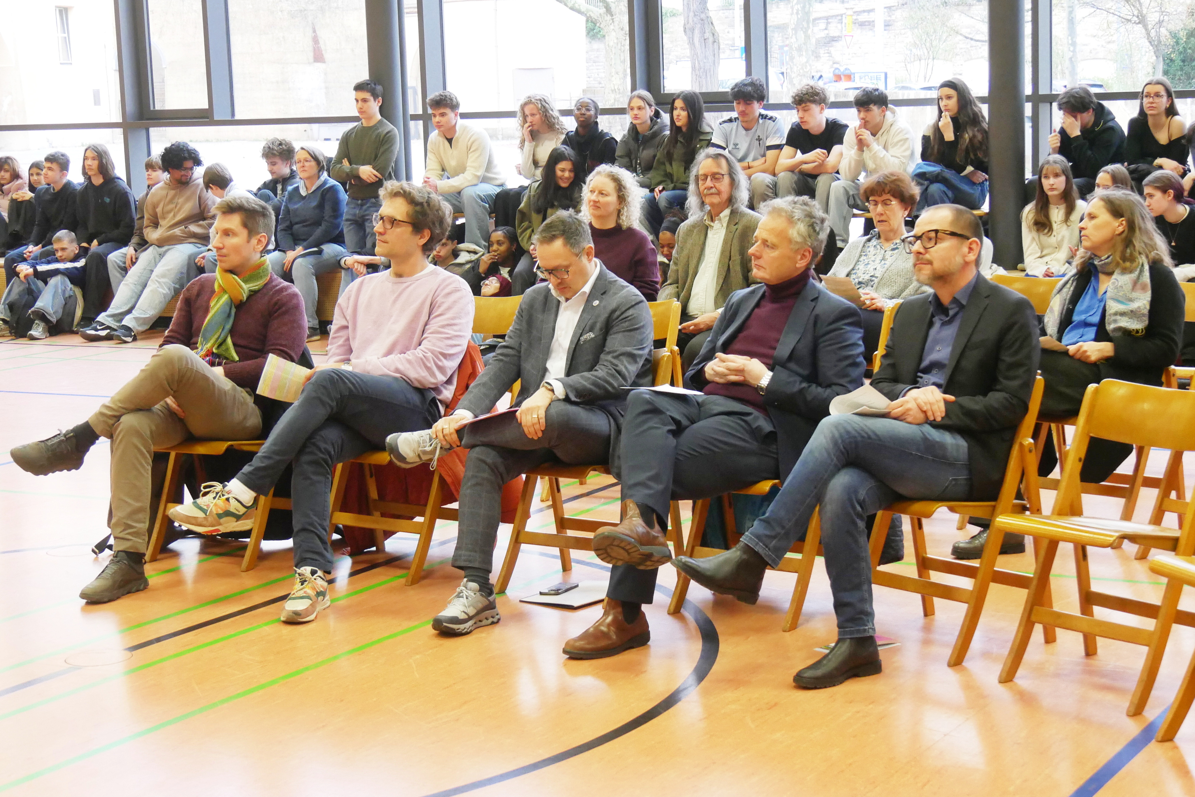 Sitzende Gruppe mit neutralen Gesichtern in heller Turnhalle, bunte Stuehle