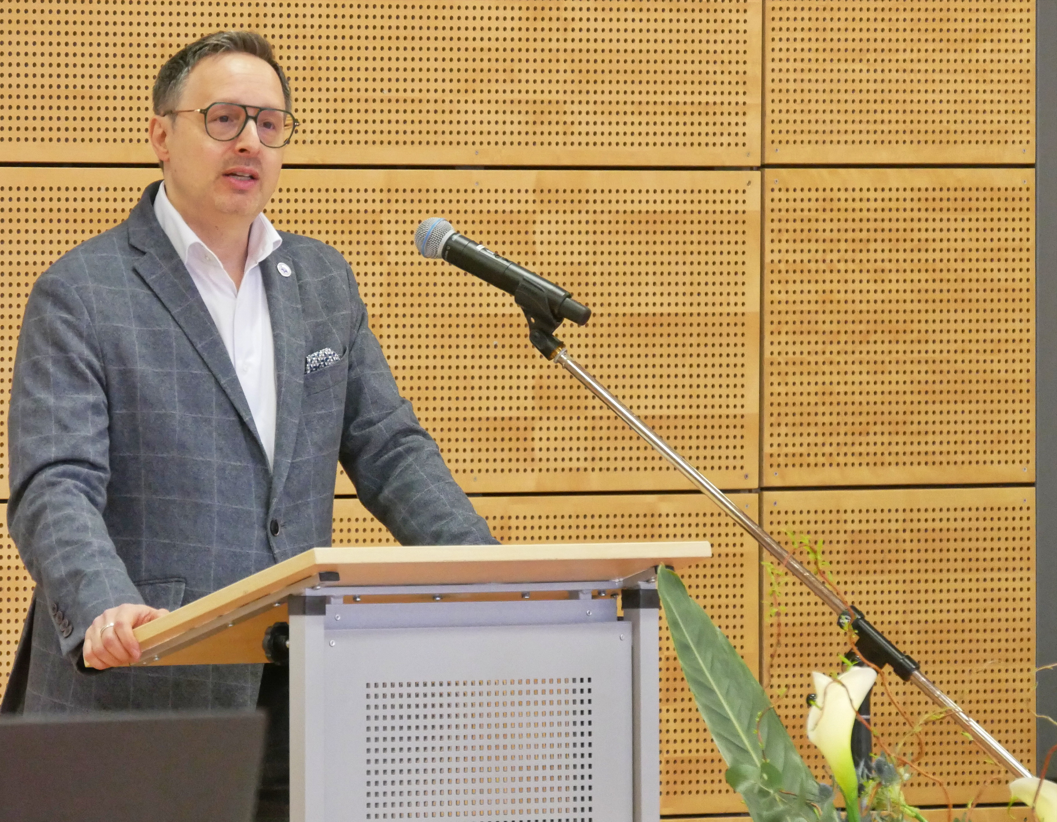 Mann mit neutralem Ausdruck im grauen Sakko am Rednerpult mit Mikrofon
