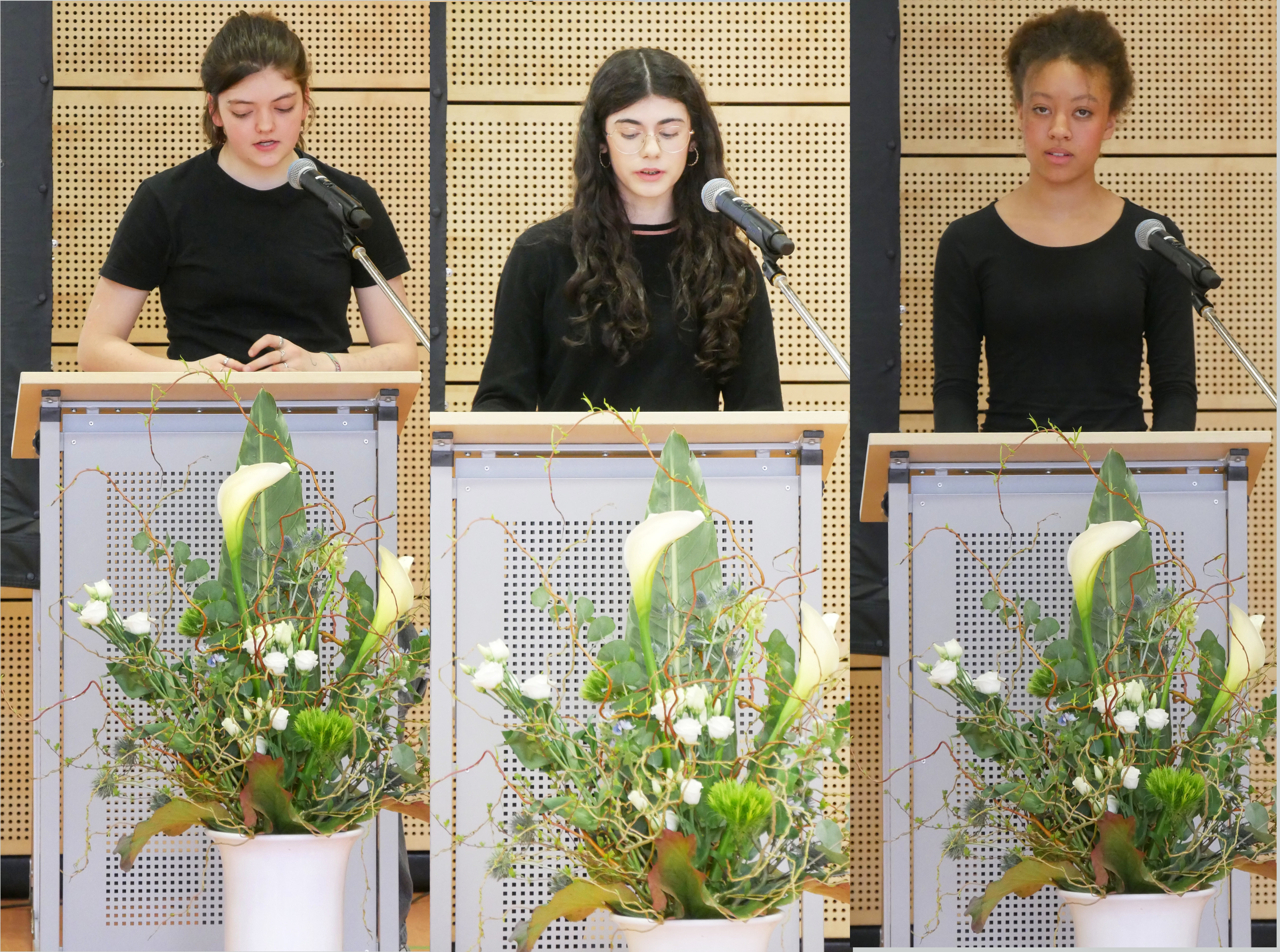 Drei Personen ernst am Rednerpult mit Mikrofon und gruenem Blumenarrangement