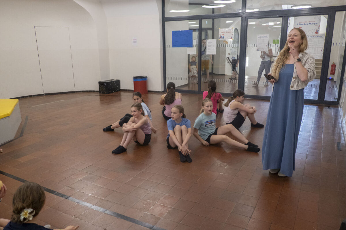 Frau im blauen Kleid lächelt, Kinder sitzen im braunen Flur, Glaswand