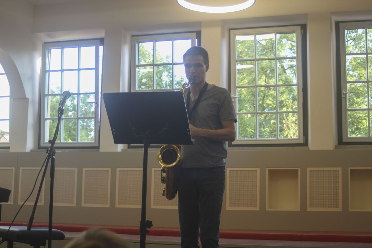 Mann mit neutralem Ausdruck spielt goldenes Saxofon im grauen Outfit am Notenstaender
