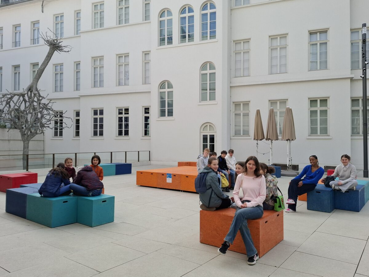 Menschen sitzen entspannt auf bunten Würfeln im hellen Hof mit Skulptur
