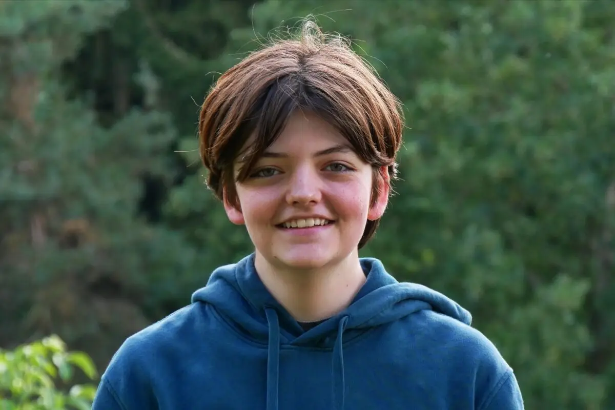 Lachender Jugendlicher mit braunem Haar im blauen Hoodie vor Gruen[0m