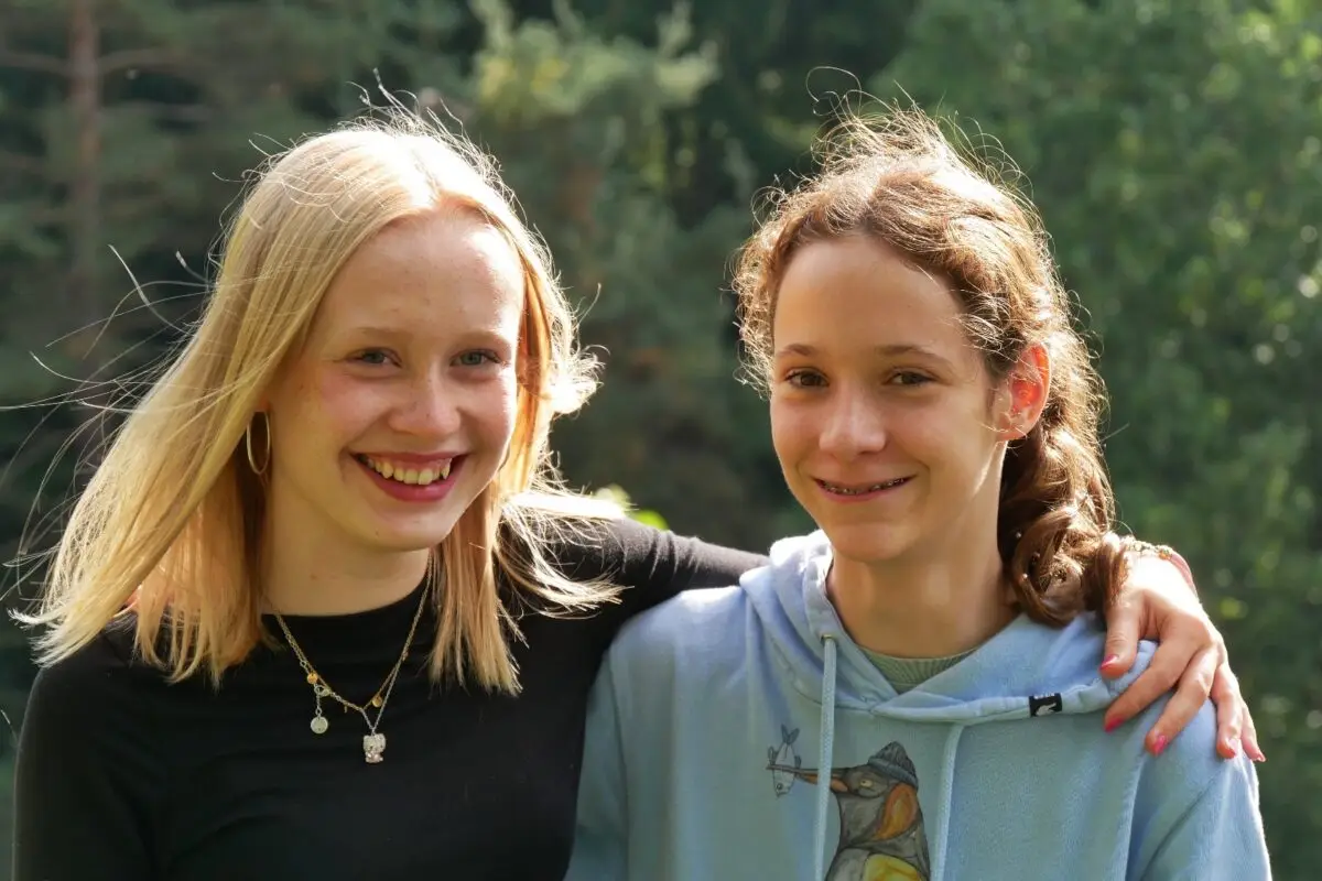 Zwei junge Frauen lächeln, blond in Schwarz und brünett in Blau, Arm um Schulter