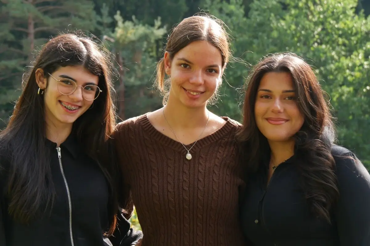 Drei laechelnde Frauen mit dunklem Haar, brauner Pulli, Brille, im Gruenen