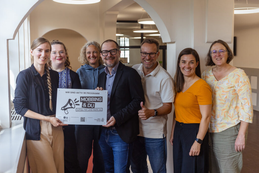 Sieben laechelnde Personen halten ein Schild zum Programm Mobbing und Du