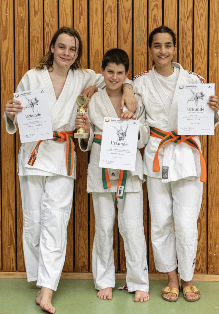 Drei laechelnde Judokas in weissen Anzuegen halten stolz Urkunden und Pokal