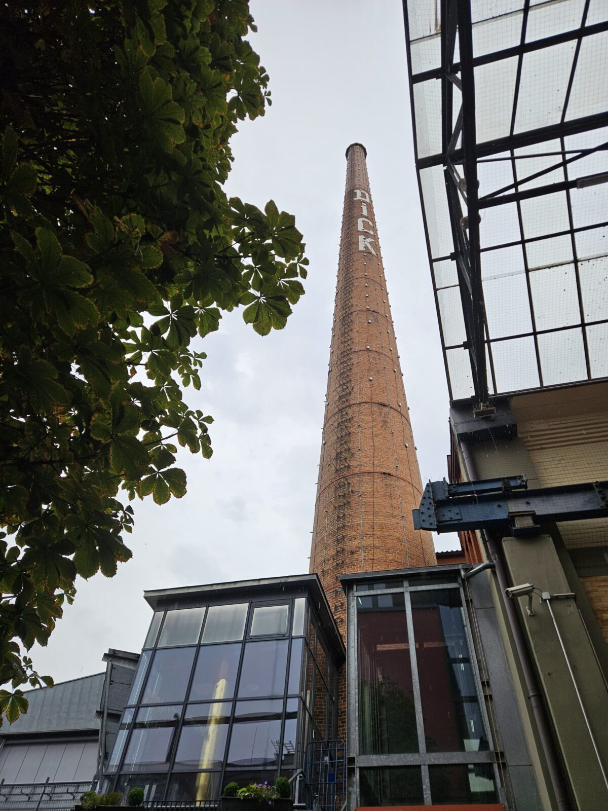 Hoher Backsteinschornstein mit der Aufschrift Dick neben Industriegebaeuden und Baum
