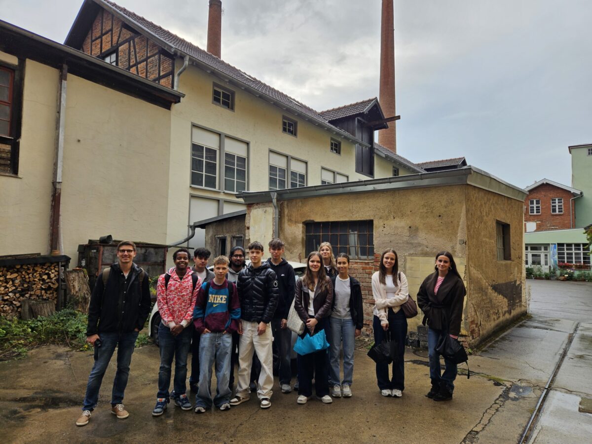 Laechelnde Gruppe junger Menschen vor alten Fabrikgebaeuden mit Schornstein