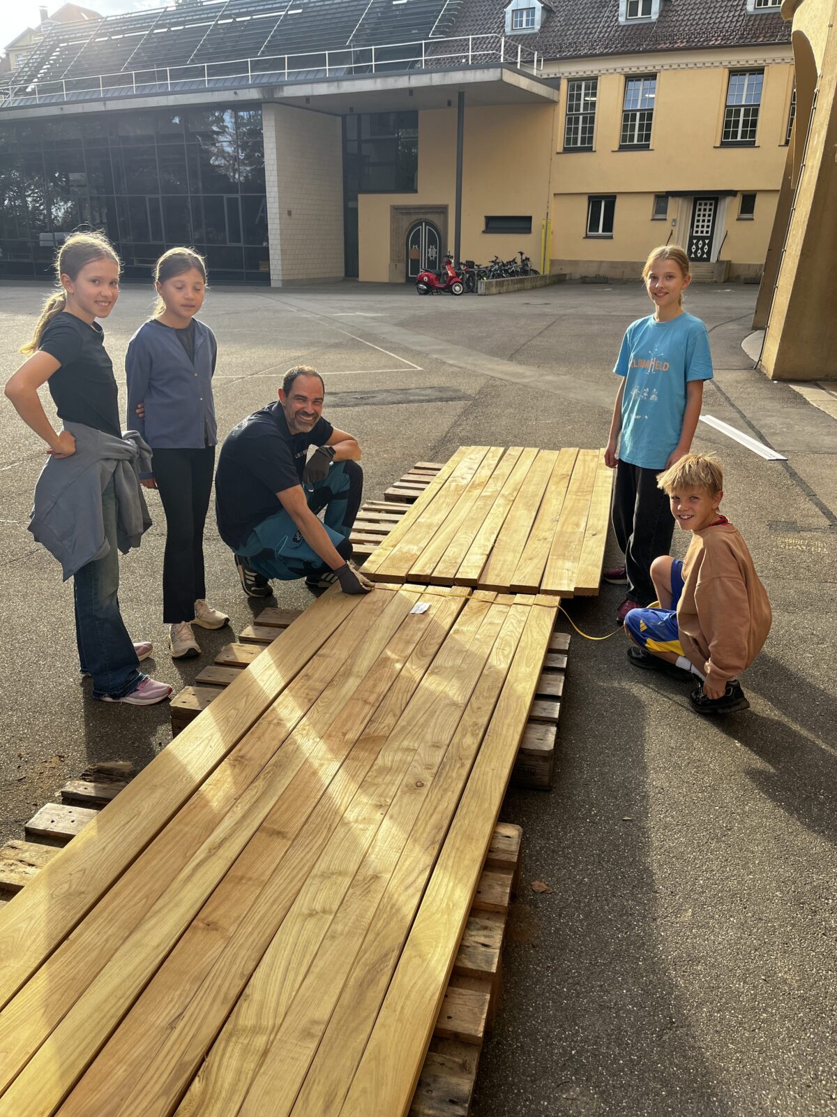 Ein laechelnder Mann und vier Kinder stehen an frischem Holz auf einem Schulhof