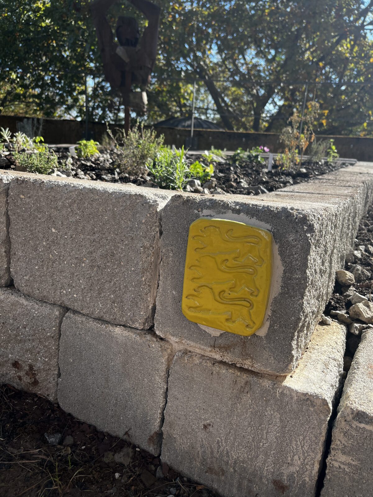 Eine gelbe Keramikkachel mit drei Loewen auf einer grauen Steinmauer im Garten