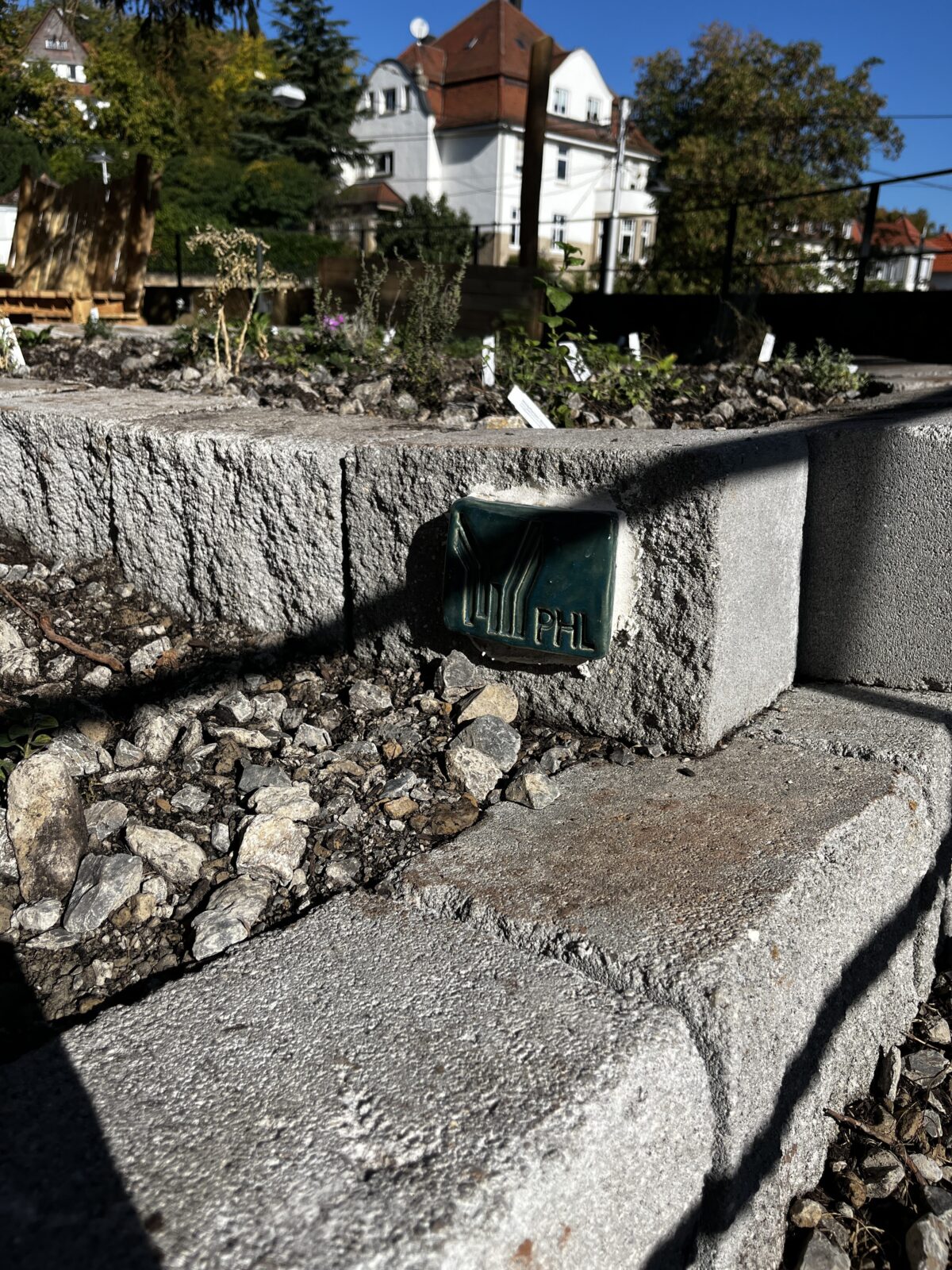 Gruene Keramikfliese mit PHL Praegung in einer Betonmauer vor weissem Haus