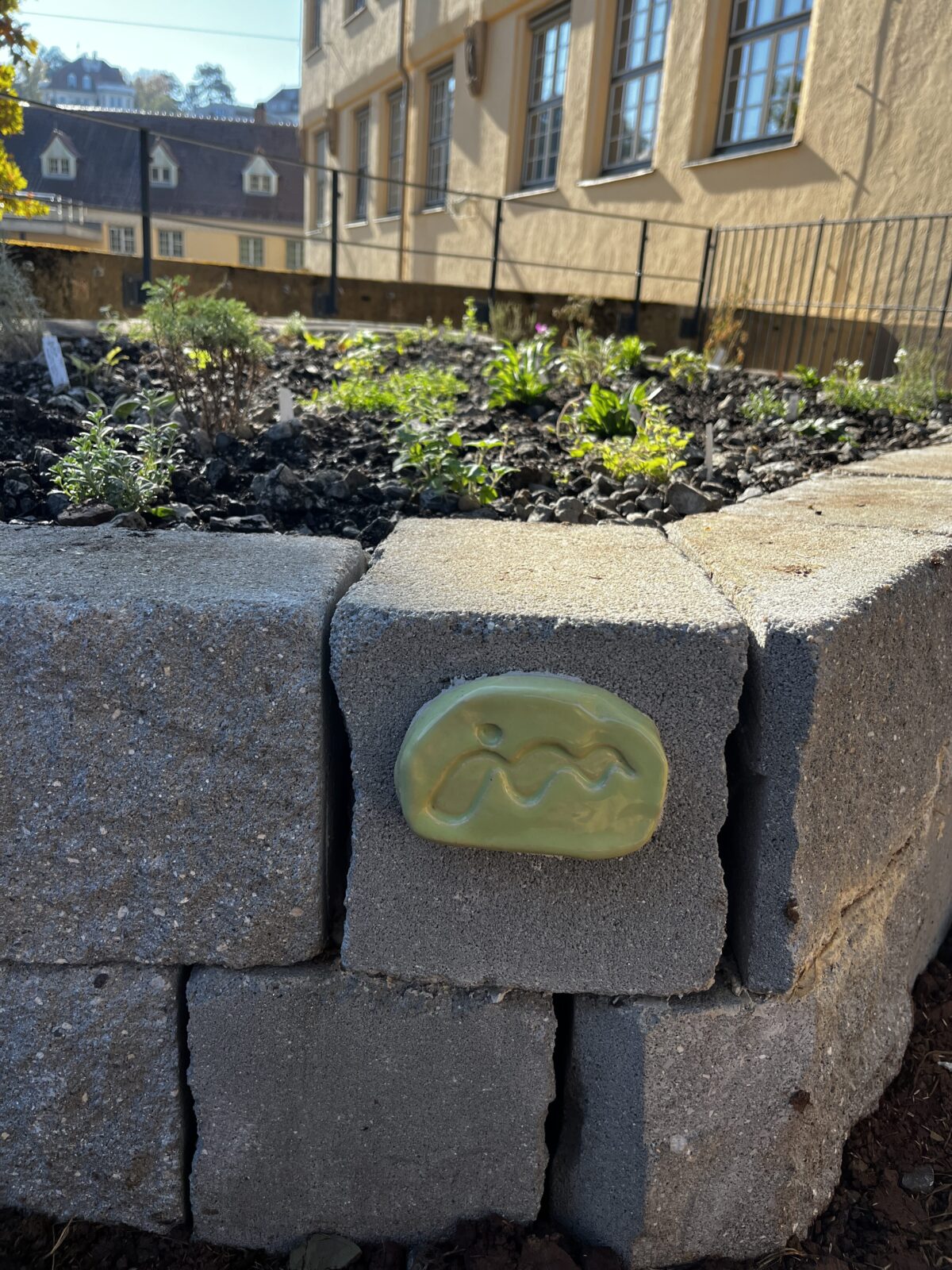 Grüne Keramikplakette mit Wellenmuster auf einer Betonmauer vor einem Beet