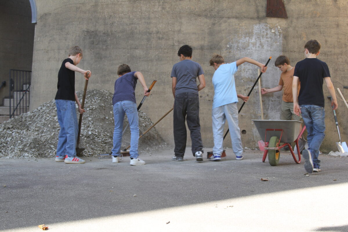 Sechs Jungen arbeiten mit Schaufeln und Schubkarre an einem Schotterhaufen